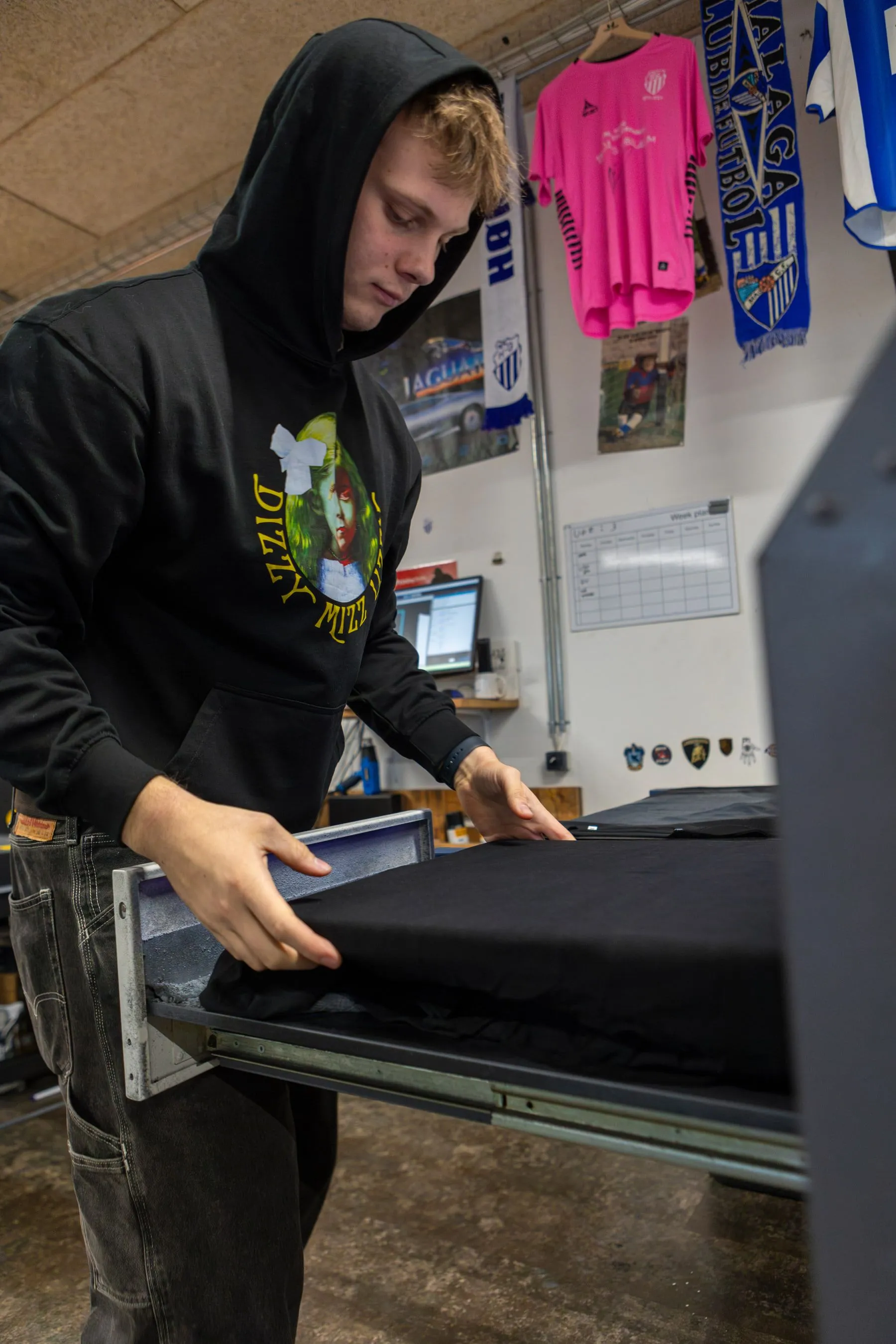 En medarbejder trykker merch ved DTG-printeren under en væg af klubtrøjer og halstørklæder.